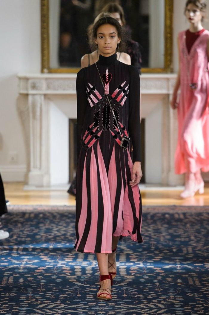 Modeshow av extravaganta klänningar - svart poloklänning med rosa pläterar och rosa tryck, långt till foten. bärs med öppna röda skor med plana sulor