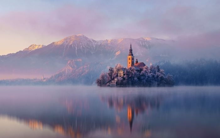 romantica scena invernale - un lago con un'isola con una chiesa e piccole case e alberi - montagne con neve e nuvole rosa