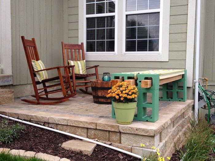casa com terraço - duas cadeiras marrons com almofadas verdes - vaso com flores de laranjeira - um banco de jardim verde feito de pedras de plantas verdes