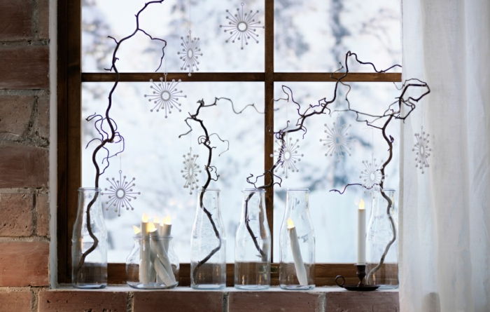 fyra torra trädgrenar placerade i glasflaskor, tre konststearinljus i glasvaser, kransar av DIY-papperssnöfling, lång vit tulledraperi, hus med tegelvägg, gammalt fönster med träfönsterramar