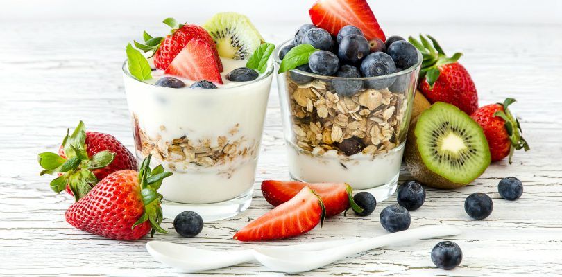 Yoghurt i glass med tre varianter av frukt - kiwi, jordbær, blåbær, servert med havregryn, hvitmalt trebord, to små porselenskjeer