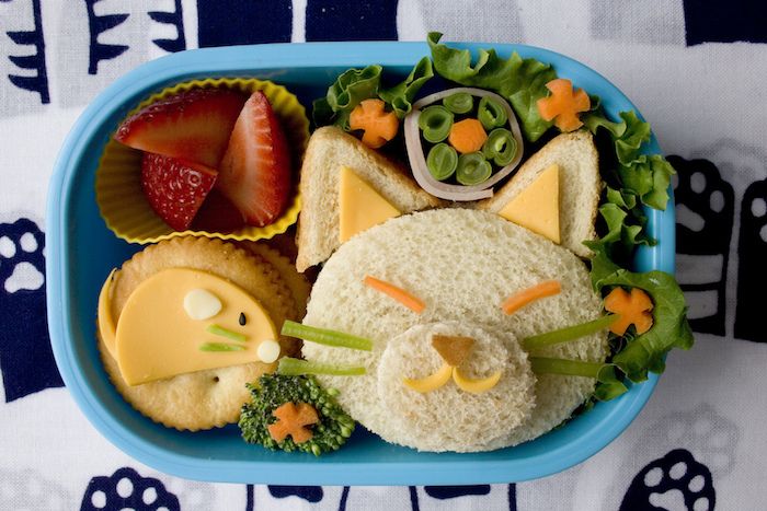 Küçük çocuklar için öğle yemeği: Bir kedinin yüzü şeklinde sandviç, yeşil biber bıçağı, doğranmış çilek, peynirli bisküviler, yayın balığı baskı masa örtüsü