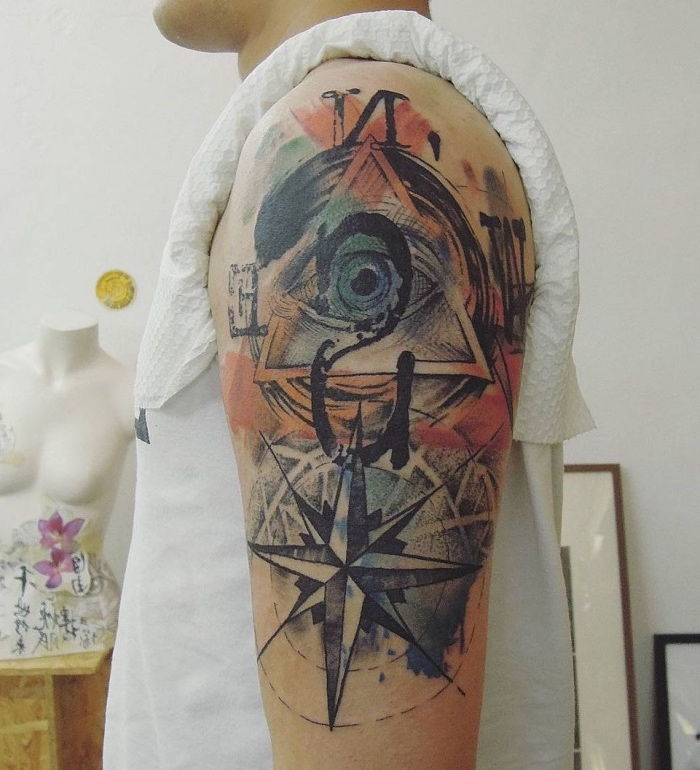 Žmogus su beprotiška spalvinga tatuiruotė ant jo peties, su dideliu mėlyna akimi ir didelis juodas kompasas