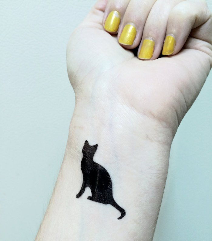 świetny pomysł na tatuaż kota na nadgarstku - tu ręka, żółty lakier do paznokci i mały czarny kot