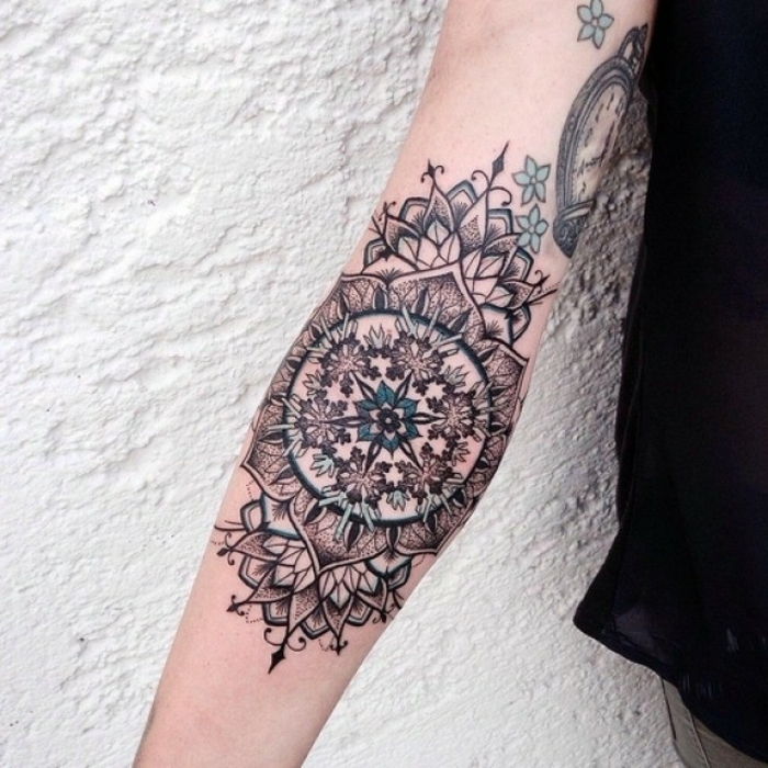 Kvinna med underarm tatuering med komplicerad design i två färger - svart och turkosblå, blå blomma i mitten, små turkosblommor på övre sidan