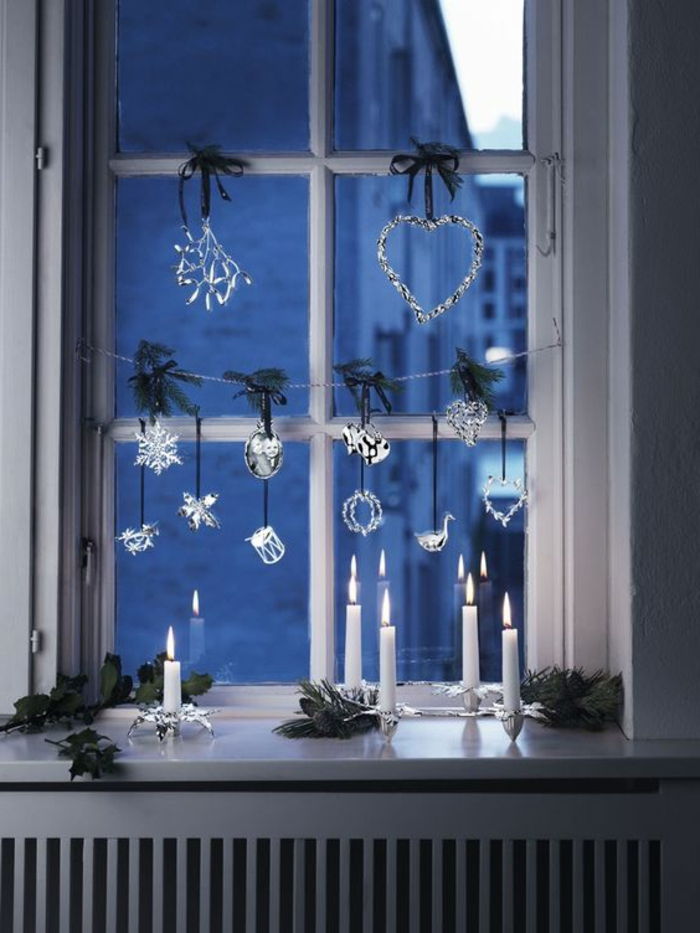 Fensterdeko for julaften - hvite stearinlys på elegante lysestaker, små biter Conifer grener, vindu krans av glass figurer, dekorativt element i form av et hjerte med små LED-lys og et grønt bånd oppvarming panel laget av tre