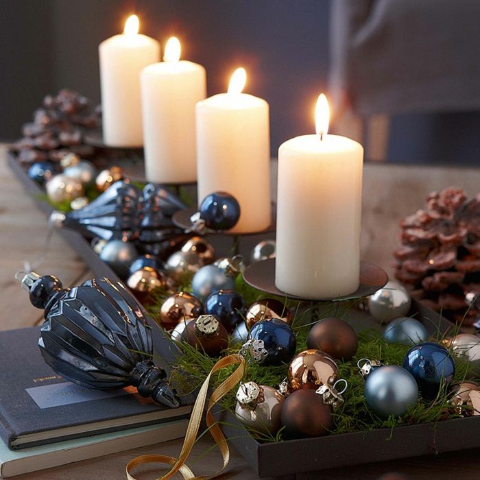 fire lys på svart lysestaker laget av rustfritt stål, små juletrekuler i forskjellige farger med glans og matt belegg, dekorativt element for juletre laget av blått, lett knuselig glass, to store kjegler på en rikt dekorert bord, to bøker - en med mørk blå og ett med lys grønn konvolutt