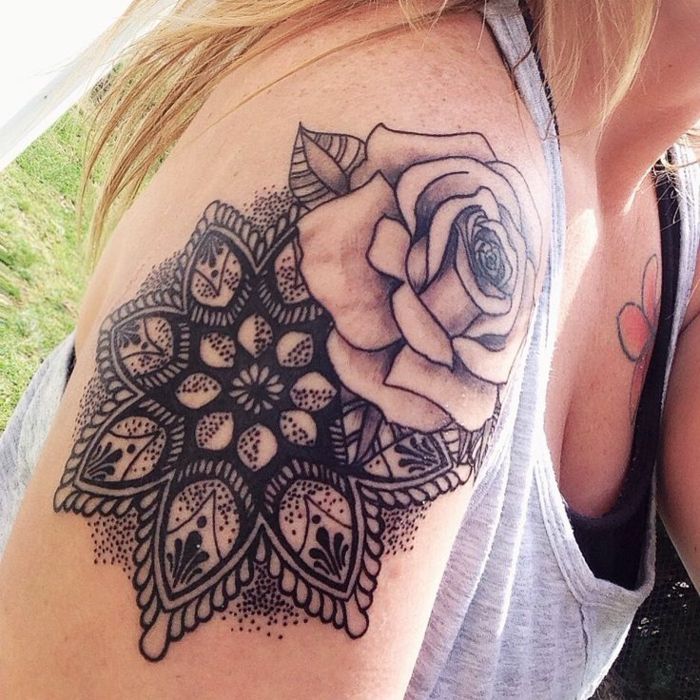 en stor grå ros med ett blad på vänster sida och en svart mandala med många svarta prickar, tatuerad på höger axel av en blond med golblonden hår, klädd i en svart topp och en blomma tatuering på vänster sida av bröstet har