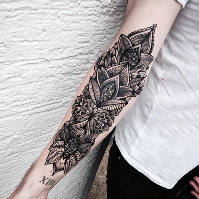 Kvinna med svart underarm tatuering på höger arm, svart tatuering med komplexa motiv, blomma och bladmotiv med många linjer, tatuering med datum, svarta byxor med vit t-shirt
