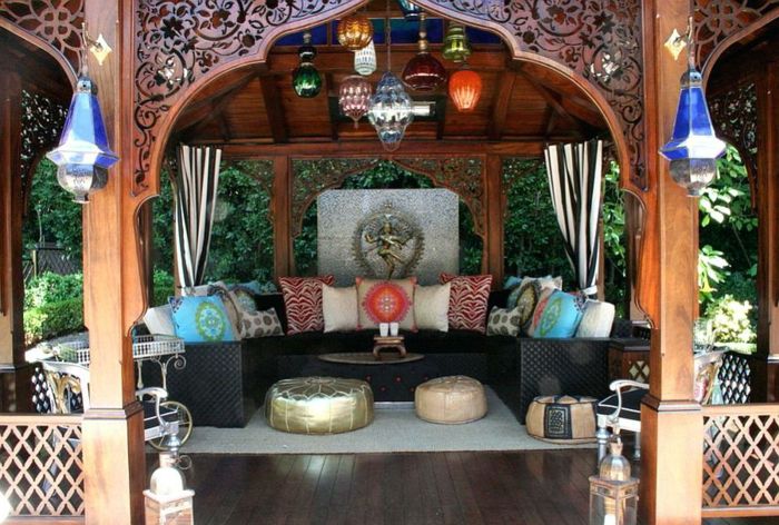 Indijski sedenje na vrtu - velika Flechtcouch v črni barvi z veliko blazine v različnih barvah, gazebo z lesene rezbarije, puder v usnje, preprogo v svetle barve, lesena tla v temne barve, dvema stranskima mize in bel naslonjač s črnim oblazinjenega sedeža, dekoracijo sveč in veliko malih stekleni lestenci