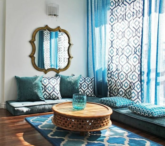 tipično maroško sedenje v odtenkih modre in bele barve, mehko vzorec preprogi v treh barvah, veliko stensko ogledalo z nepravilno obliko in pozlačeno okvir z zastarelo podobo, majhne stenske svetilke o nizki okrogli mizi kave iz lesa, modre steklene vaze z okrogle oblike, tri dolge pol prosojne zavese v blue