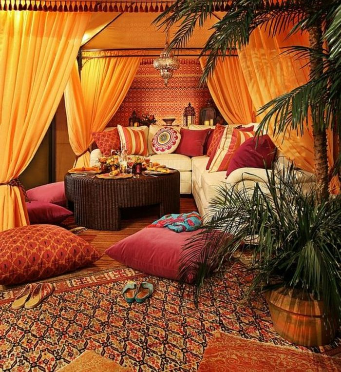 Bivalni prostor z Baldahine z oranžnimi zavesami, bel kavč iz usnja, rdeči kavč blazine v untershiedlichen velikostih, okrogle Flechttisch v temno rjave barve, velik vzorec preproge z cvetlični lonec na velike in majhne palme, dekorativni elementi iz kovine