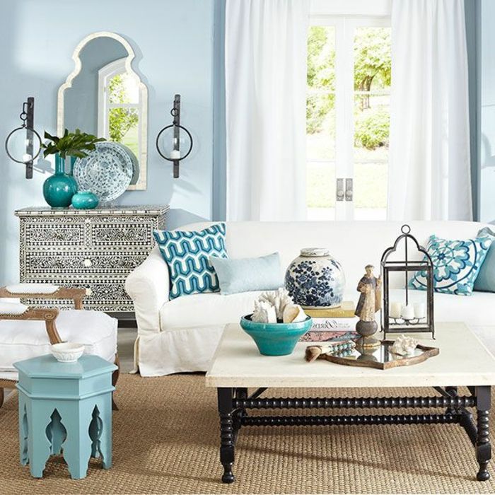 Dnevna soba s svetlo modro steno, dolge bele zavese, dekorativne ogledalo z ozkim belim okvirjem, dekorativne plošče v dveh barvah, kava miza narejena iz lesa, pobarvan v svetlo modro, belo mizo s črnimi lesenimi nogami ahagoniholz, vzorec blazine v modri, veliko dekorativnih elementov na mizi