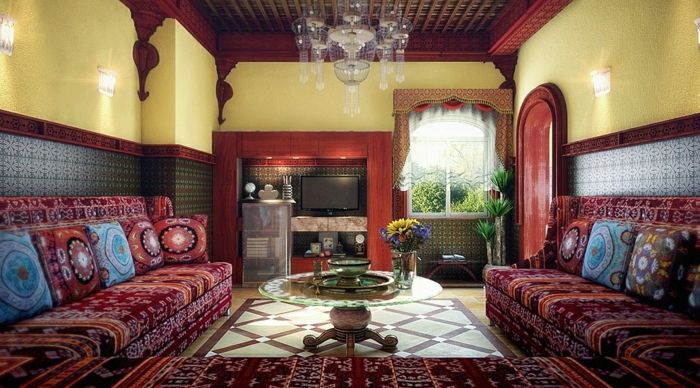 Maroški Wohhnzimmer flat TV v stenski niši, rumene stene z vzorčastim ozadje v modri, posredno razsvetljavo v steno v dnevni sobi, lestenec kristalov, okroglo stekleno mizo z nizkimi lesenimi nogami, kavč z maroškimi tkanine