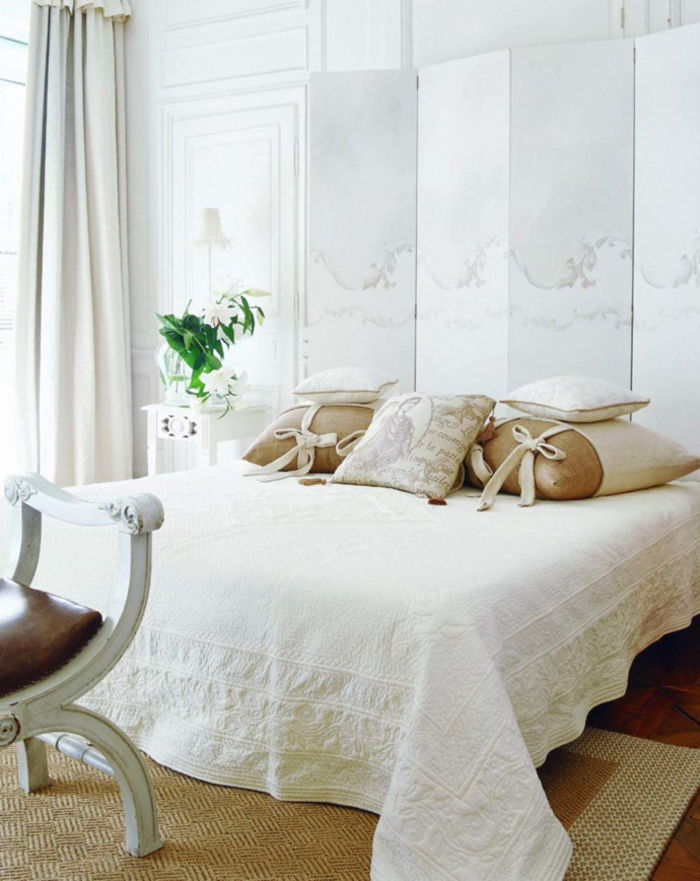 Camera da letto vintage in bianco, molti cuscini decorativi, vaso di fiori, lampada da comodino